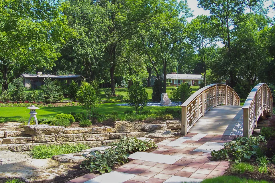 Wood bridge and stone-lined garden channel with paver path, lantern, clipped planting, and lawn.